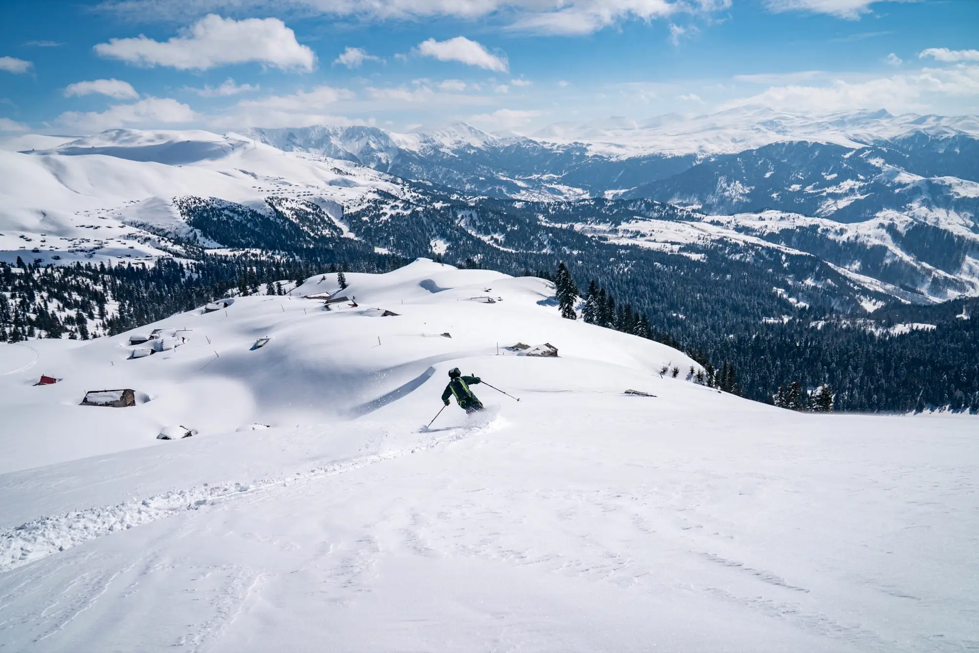 Skiër daalt af over een open helling, met houten hutten en bergen in de verte